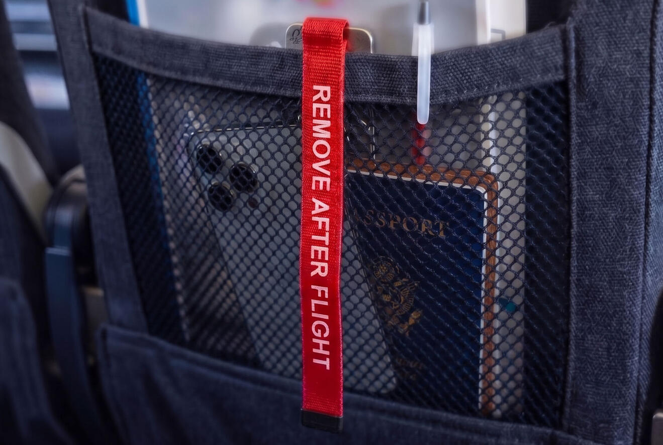 A close-up view from a passenger's perspective looking down into the fabric seatback pocket of an aircraft seat. The red 'REMOVE AFTER FLIGHT' streamer tag dangles visibly from the top edge of the pocket. Travel documents and a pen are partially visible in
